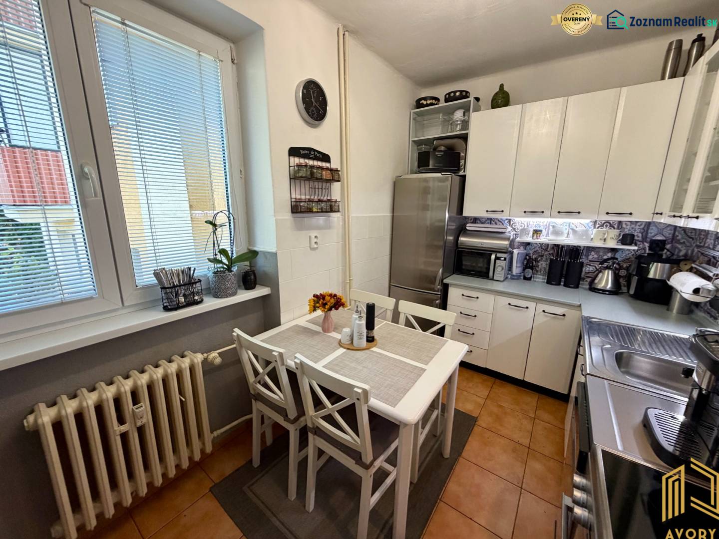 Konyha egy háromszobás lakásban asztallal, hűtőszekrénnyel és háztartási gépekkel. Fehér szekrények és csempézett padló.