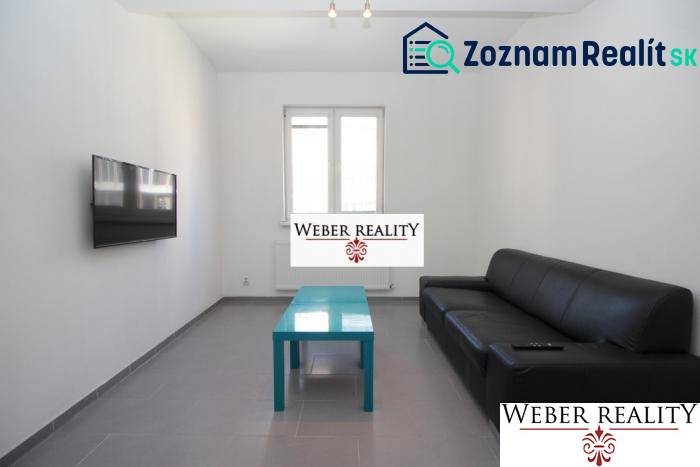 Nappali egy kétszobás lakásban fekete kanapéval, falra szerelt televízióval és alacsony asztallal.