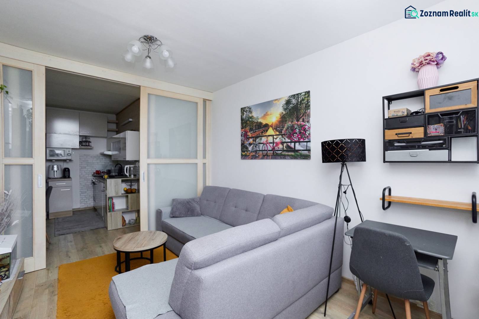 Nappali egy kétszobás lakásban, fa dekoros padlóval, szürke kanapéval és konyhához való hozzáféréssel.