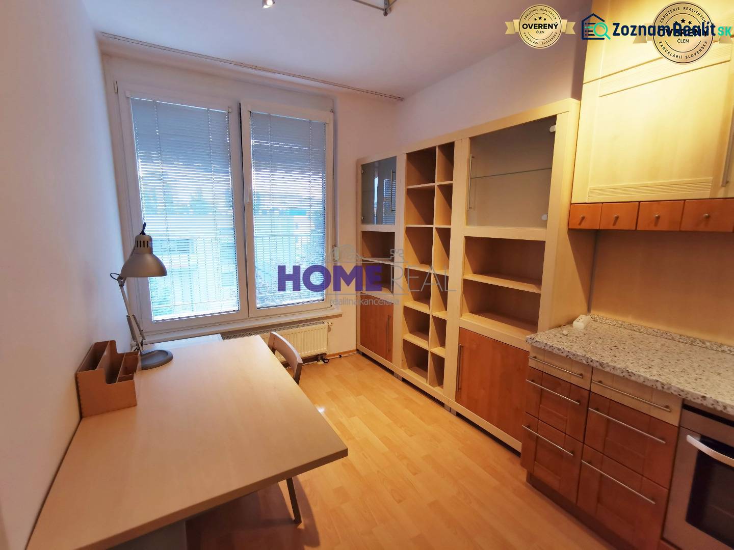 Dolgozószoba egy kétszobás lakásban, fa mintázatú padlóval, asztallal és könyvespolccal.