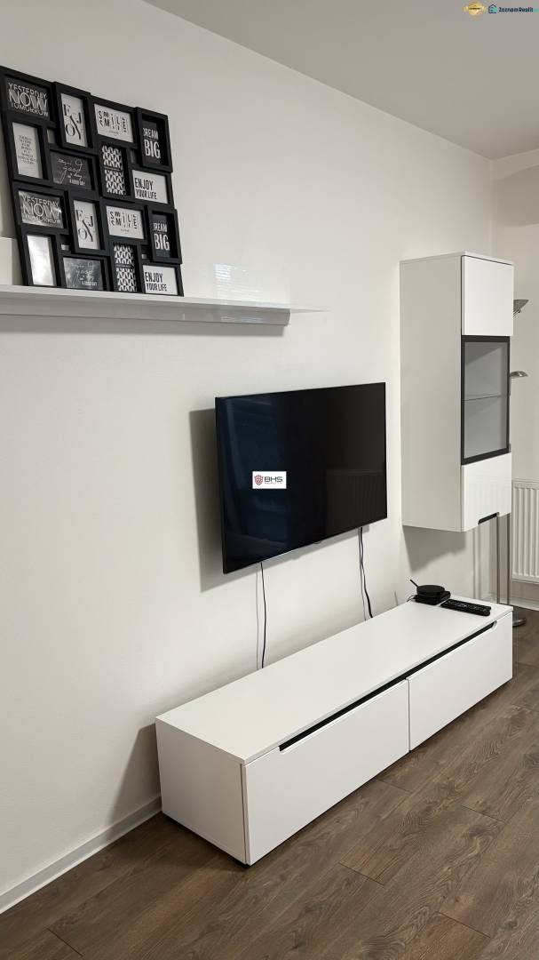 Televízió a falon, fehér komód, képek és fa mintázatú padló az egyszobás lakásban.