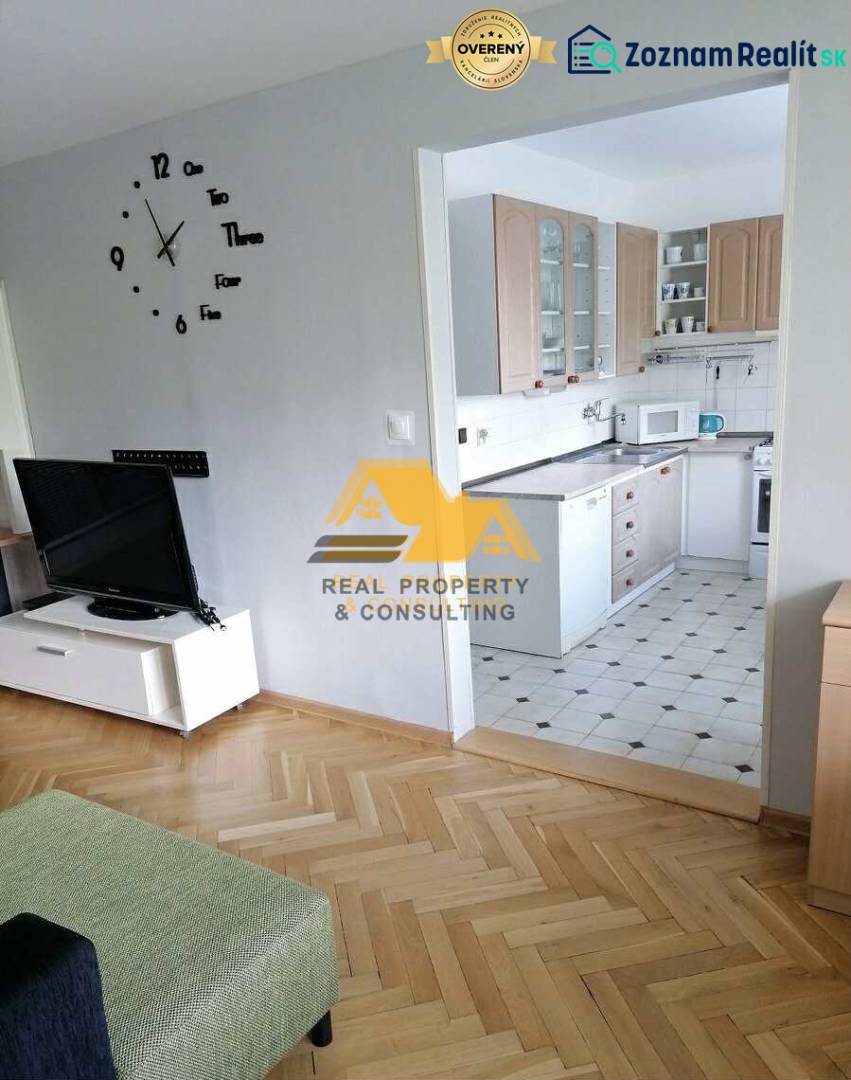 Nappali egy háromszobás lakásban, fa mintázatú padlóval és háttérben konyhával.