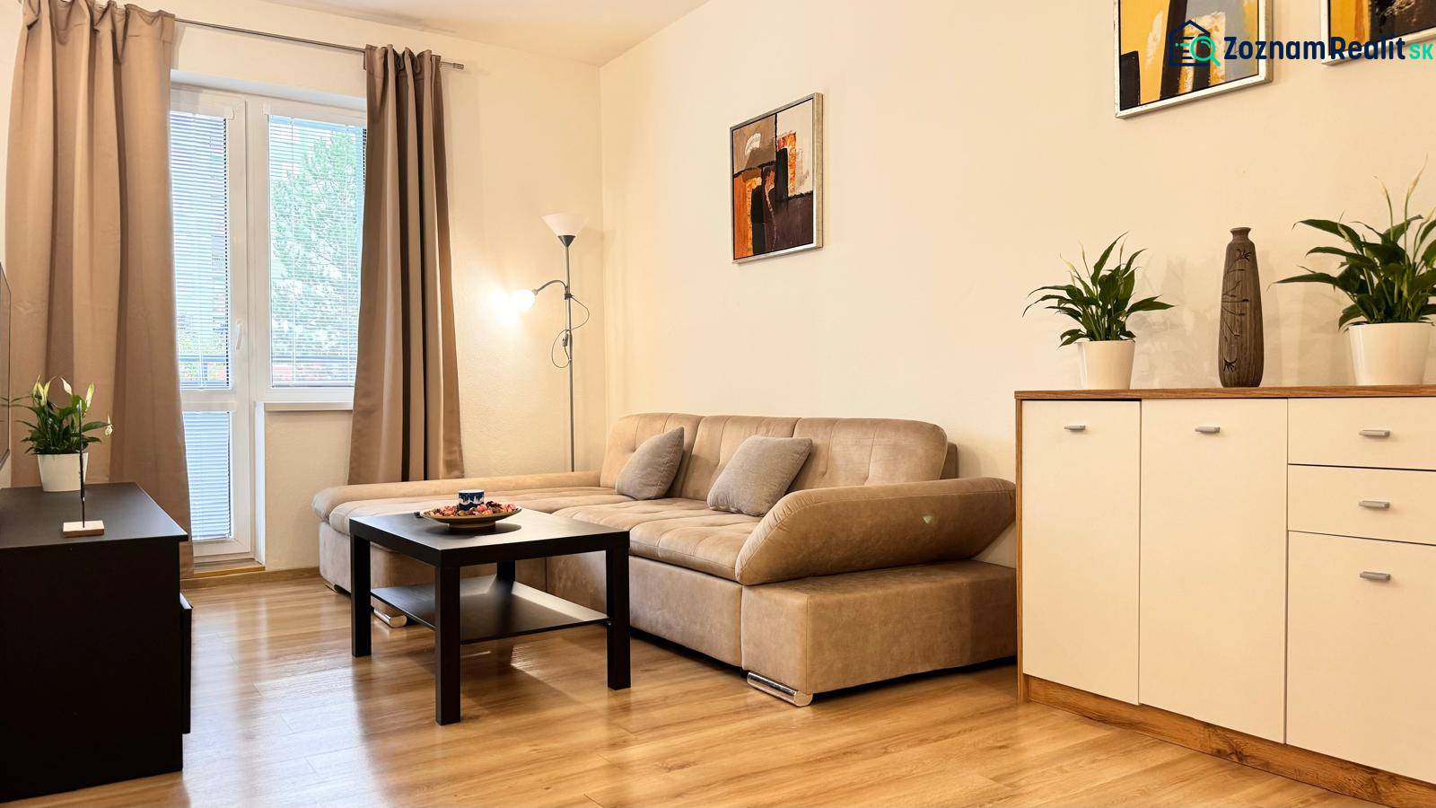 Nappali egy kétszobás lakásban bézs kanapéval, fa mintázatú padlóval.