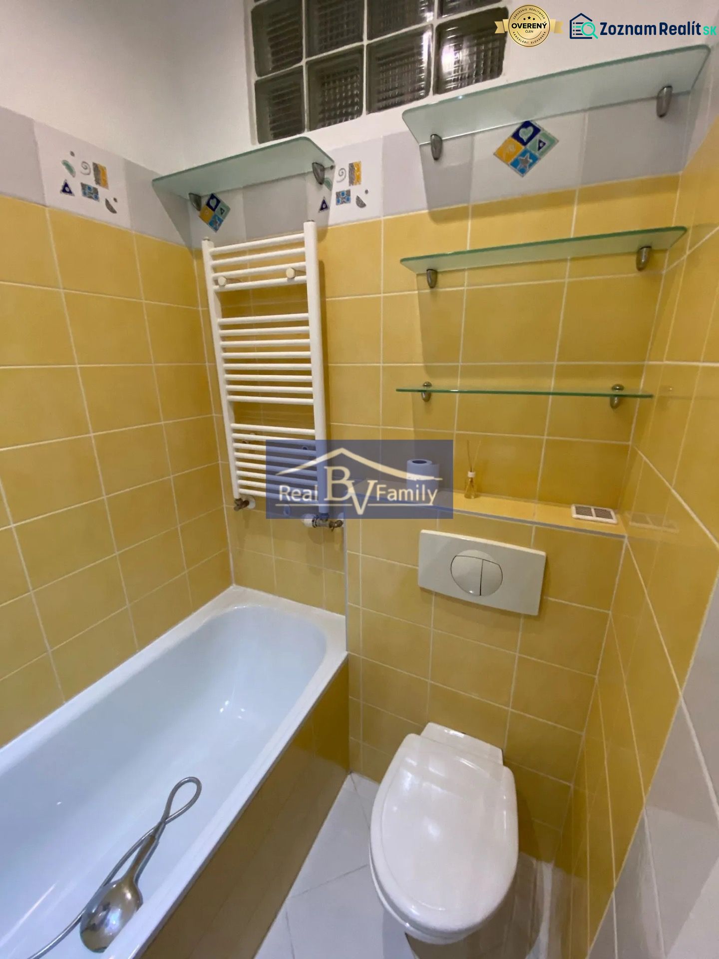 Fürdőszoba sárga csempékkel, káddal, WC-vel és üvegpolcokkal egy egyszobás lakásban.