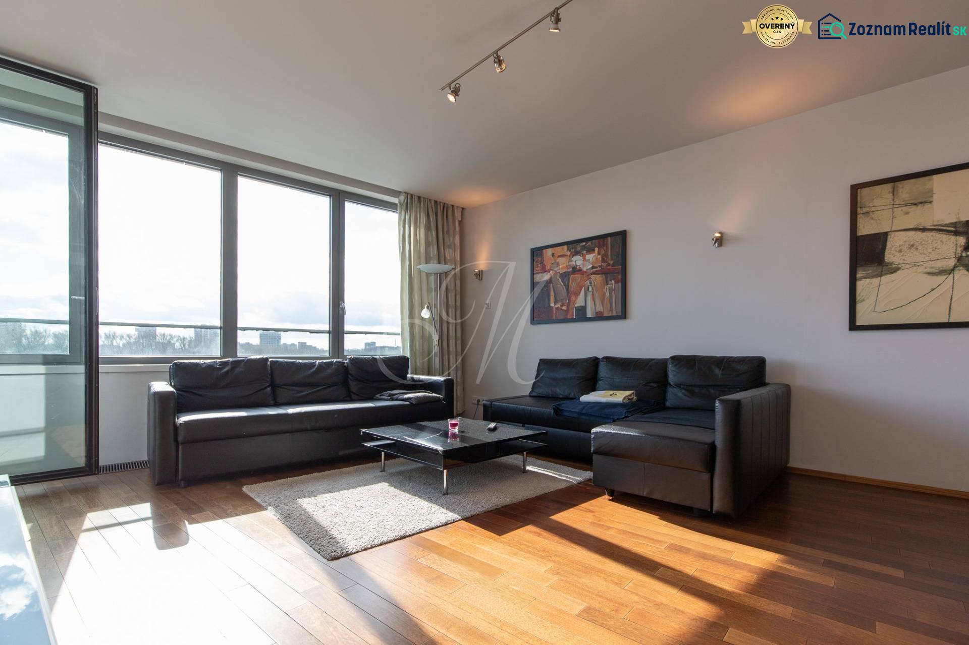 Nappali egy kétszobás lakásban fekete kanapéval, fa hatású padlóval és nagy ablakokkal.