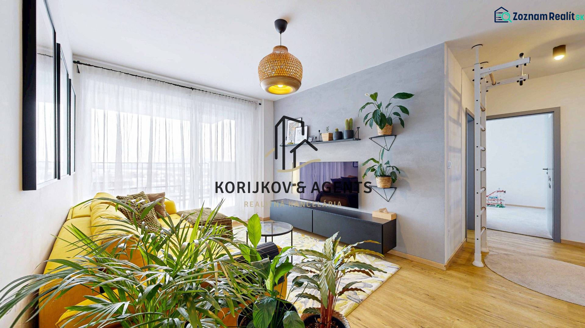 Nappali egy háromszobás lakásban nagy sárga kanapéval és növényekkel, fa dekoros padlóval.