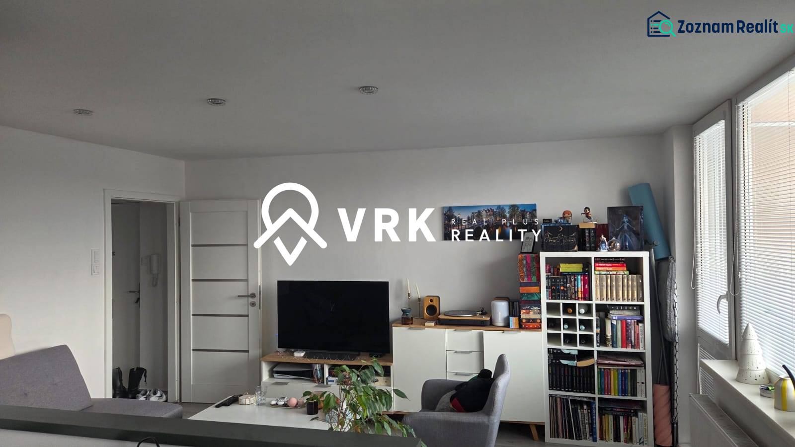 Nappali egy háromszobás lakásban könyvespolccal, televízióval és világos bútorokkal.