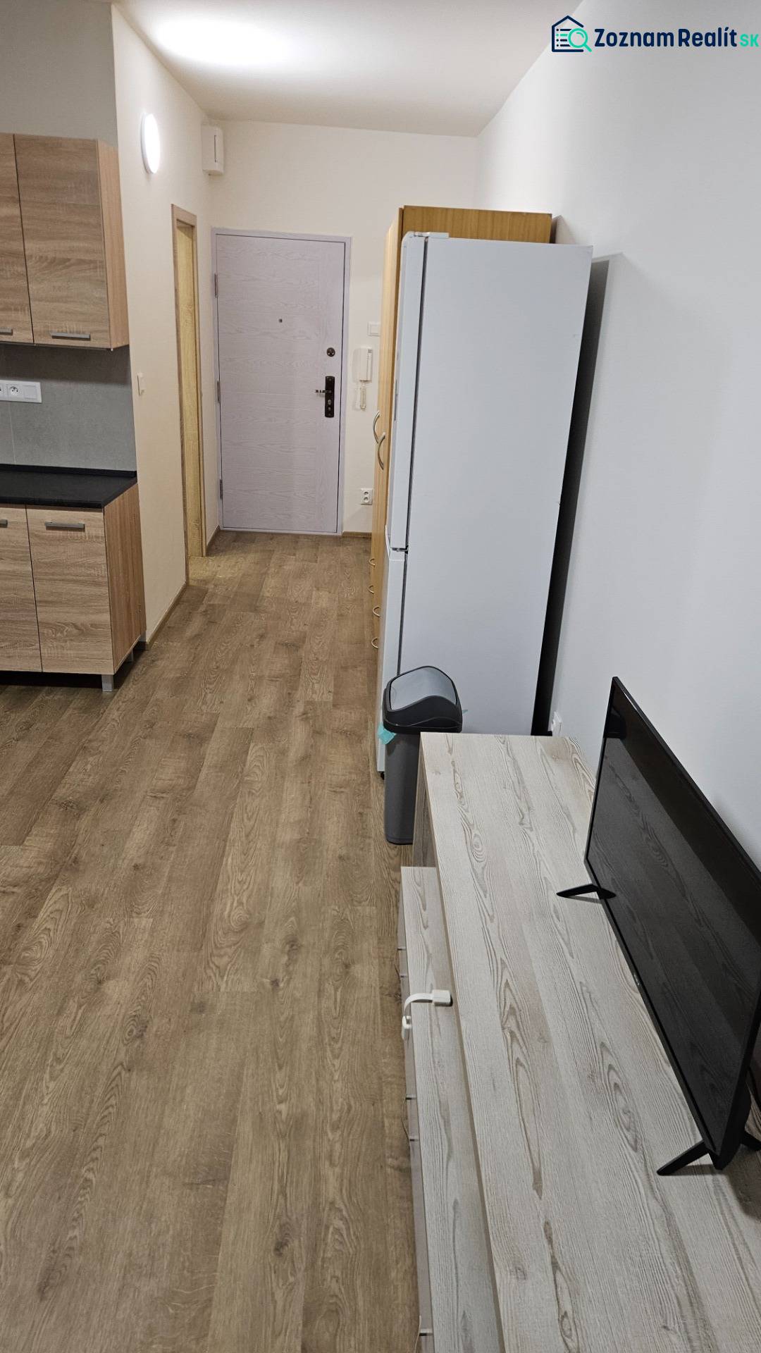 Garzonlakás fa mintázatú padlóval, konyhabútorral, hűtőszekrénnyel és televízióval.