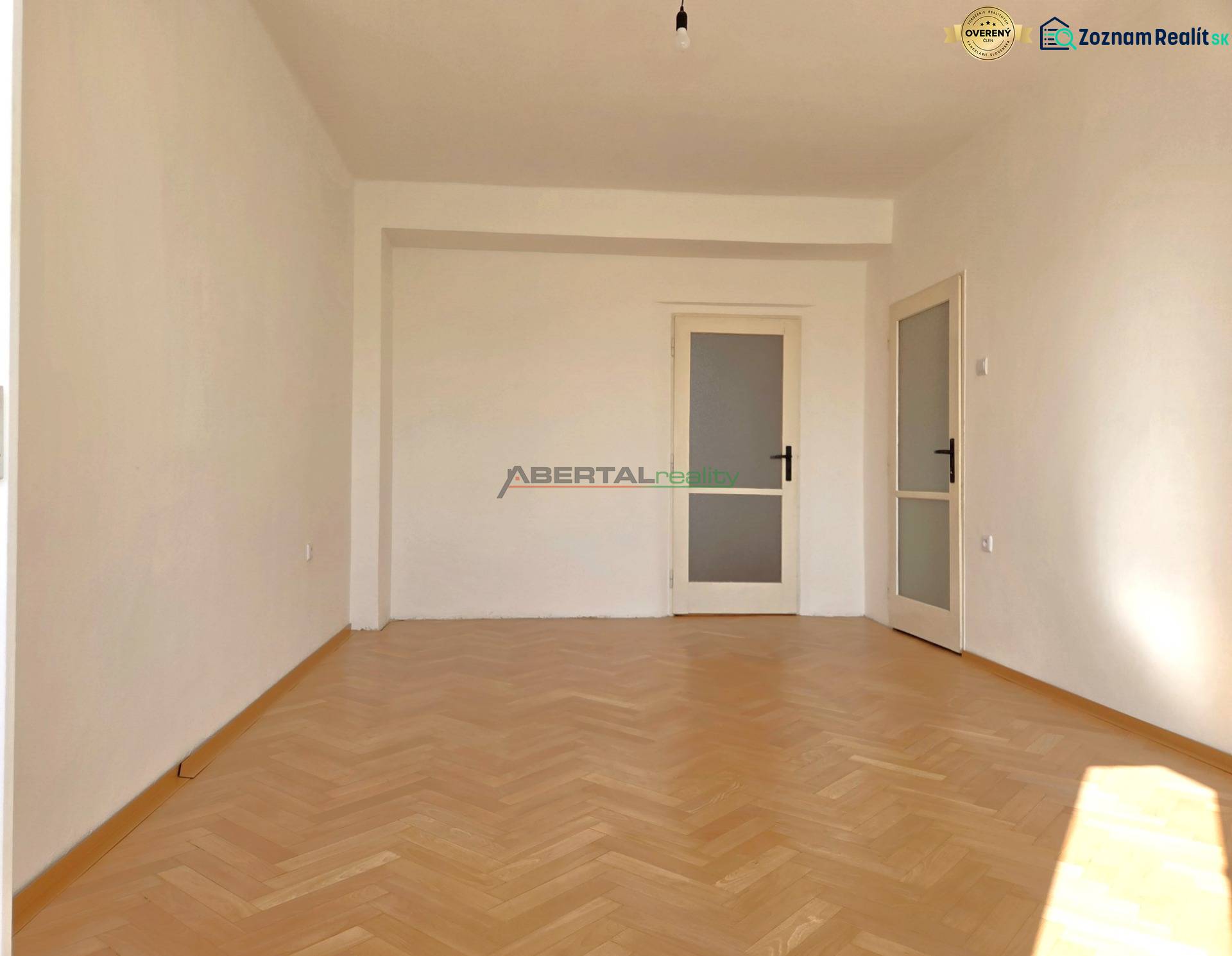 Egy szoba a háromszobás lakásban fa dekorációjú padlóval és fehér falakkal rendelkezik.