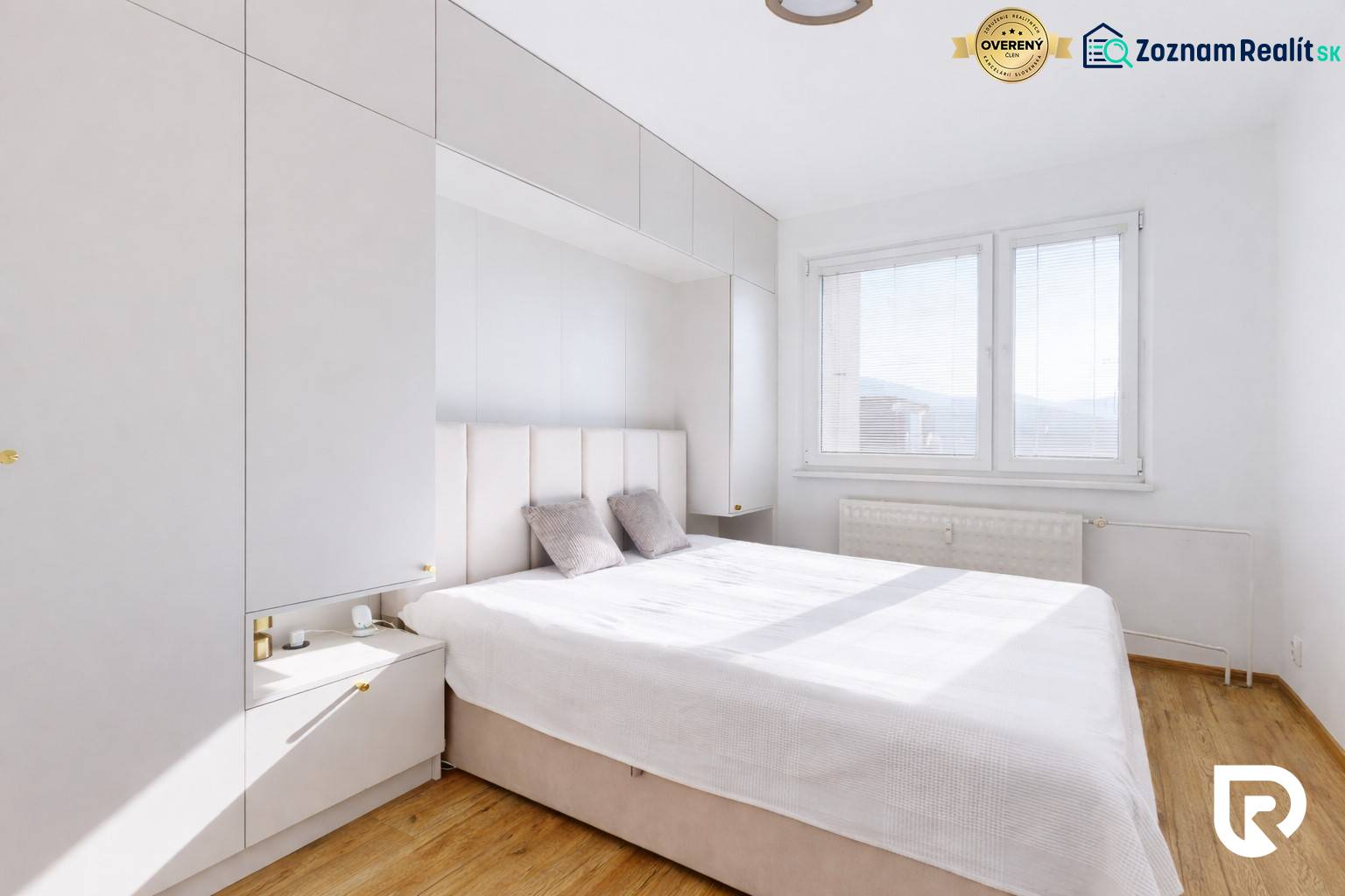 Hálószoba egy háromszobás lakásban, fa dekor padlóval, fehér bútorokkal és nagy ablakkal.