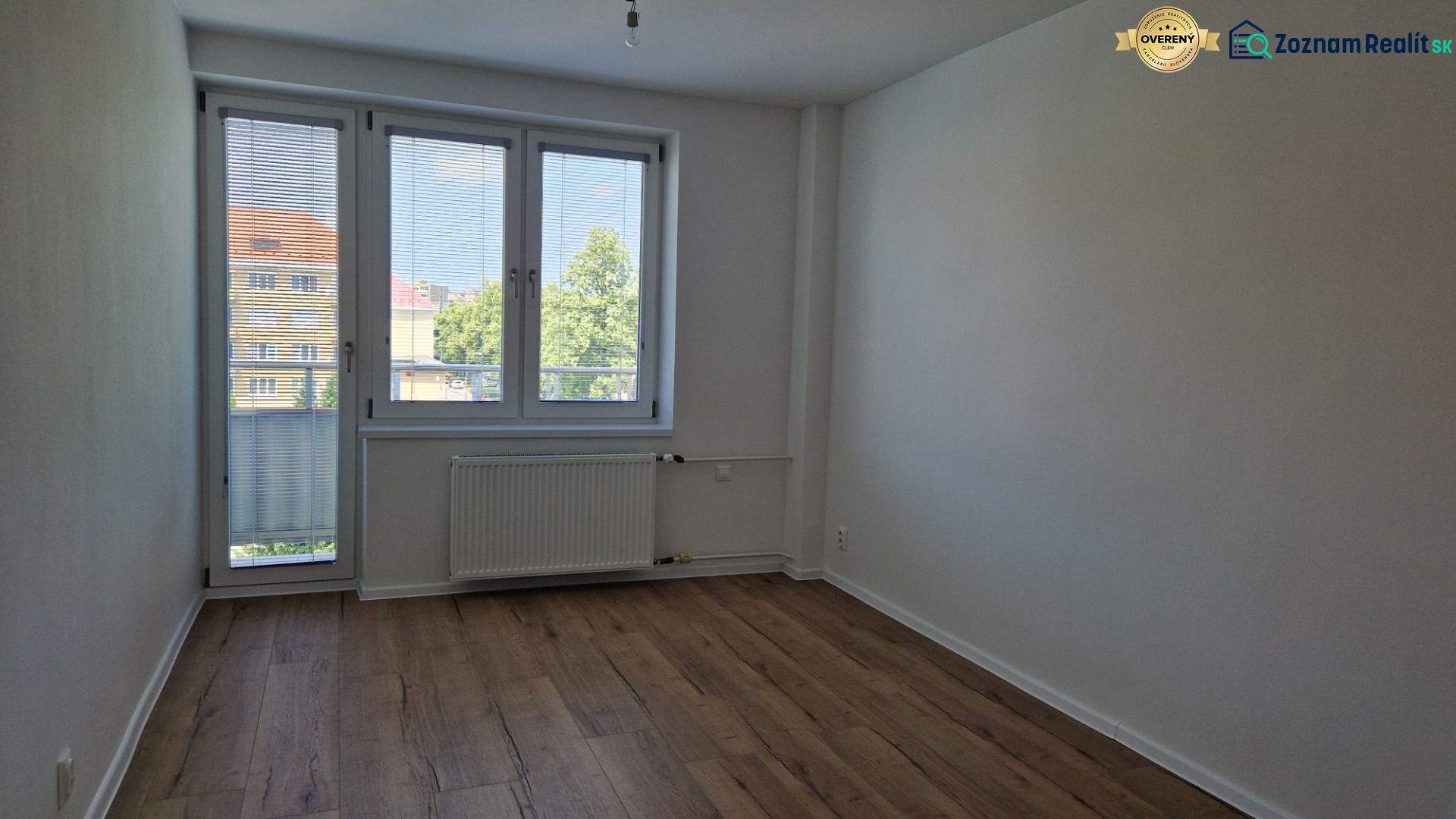 Szoba egy háromszobás lakásban fa dekor padlóval, nagy ablakkal és radiátorral.