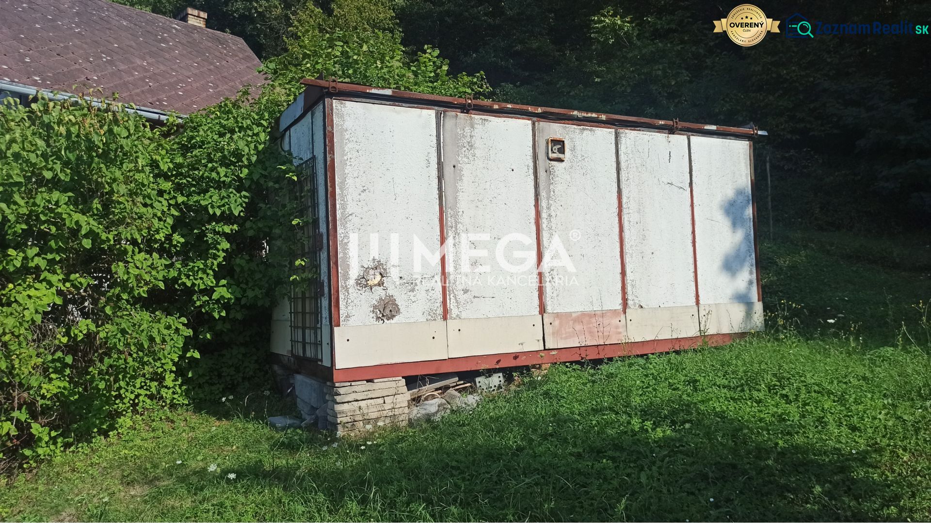 Egy régi raktár az utcai kertben Humennóban, zöldnövényzettel és növekvő fűvel körülvéve.