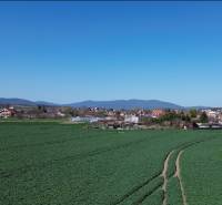 Lúky a záhrady s vidieckym zázemím, na pozadí hory a budovy pod modrou oblohou.