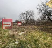 Fűvel és fákkal borított telek a Partizánska utcában, a háttérben raktárak és csarnokok.