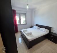 Hálószoba egy üdülő apartmanban, franciaággyal, éjjeliszekrényekkel és piros függönyös ablakkal.