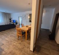 Étkezőasztal fa székekkel egy kétszobás lakásban. Része egy kanapé és egy televízió.