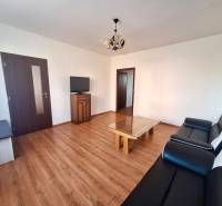 Nappali egy háromszobás lakásban, fa mintázatú padlóval, fekete kanapéval és televízióval.