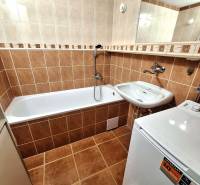 Fürdőszoba káddal egy háromszobás lakásban, barna csempék, mosdó és mosógép.