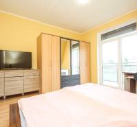 Hálószoba egy 4 szobás lakásban szekrényekkel, komóddal, TV-vel és fa mintázatú padlóval.