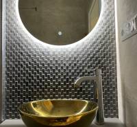 Kerek megvilágított tükör és arany mosdókagyló egy háromszobás lakásban.