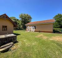 Új Szádok családi házának udvara két épülettel és kúttal, zölddel körülvéve.