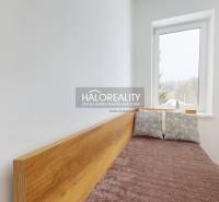 Egyszerű ágy párnákkal, fa mintázatú padló, világos szoba, nagy ablak.