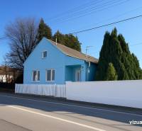 Kék családi ház Kunovban, Szenicén, a főút mellett, zöld fákkal körülvéve.