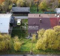 Családi ház Sirkben bádogtetővel, parkoló autóval és környező fákkal.