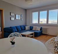 Nappali háromszobás lakásban kanapéval, fehér asztallal és fa dekorációjú padlóval.