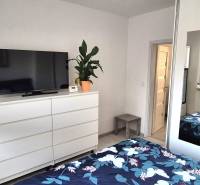 Hálószoba egy kétszobás lakásban fehér bútorokkal, tükörrel és komódon lévő televízióval.