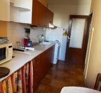 Konyha egy háromszobás lakásban, fa mintázatú padlóval, fehér szekrényekkel, étkezőasztallal.