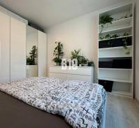 Világos hálószoba egy kétszobás lakásban, nagy szekrénnyel és polcokon lévő növényekkel.