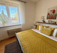 Hálószoba nagy ablakkal, sárga párnákkal és fa dekorációjú padlóval egy háromszobás lakásban.