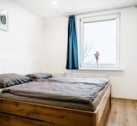 Hálószoba fa mintás padlóval és ággyal egy kétszobás lakásban, kék függöny az ablakon.