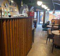 Vendéglátóhely belső tere fa bárral és asztalokkal. Dekoratív világítás is látható.