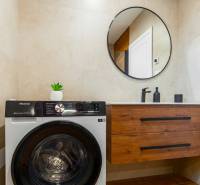Fürdőszoba egy egyszobás lakásban mosógéppel, kör alakú tükörrel és szekrénnyel.