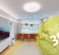 Nappali egy háromszobás lakásban, televízióval, kanapéval és fa padlódekorációval.