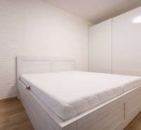 Világos hálószoba fehér ággyal, beépített szekrénnyel, fa dekorációjú padlóval egy kétszobás lakásban.