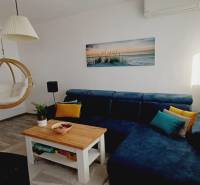 Nappali kék kanapéval, hintaszékkel, könyvespolccal és fa mintázatú padlóval.