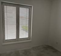 Szoba egy családi házban nagy ablakkal és redőnyökkel, fehér falak, szürke padló.