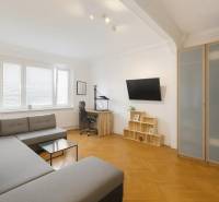 Nappali egy háromszobás lakásban sarokkanapéval, íróasztallal és fa mintázatú padlóval.