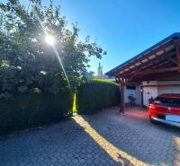 Családi ház Negyeden egy piros autóval a garázs előtt, napos nap és fa.