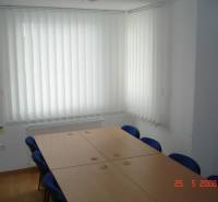 Irodai helyiség tárgyalóasztallal, kék székekkel és függőleges redőnyökkel.