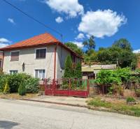 Családi ház Tachtán, piros tetővel, kerítéssel és kerttel, zöld környezetben.