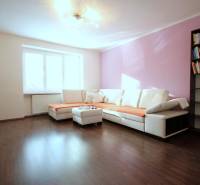 Nappali egy kétszobás lakásban sarokkanapéval, könyvespolccal és fa mintázatú padlóval.