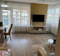 Nappali egy négy szobás lakásban, fa mintázatú padlóval és falra szerelt televízióval.