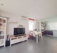 Nappali egy családi házban, televízióval, konyhasarokkal és asztallal, három székkel.