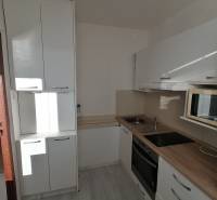 Konyha egy egyszobás lakásban, fehér szekrények, mosogató, főzőlap, mikrohullámú sütő, fa dekor padló.