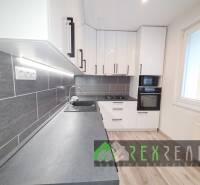 Konyha egy háromszobás lakásban fehér szekrényekkel, szürke munkalappal és fa mintázatú padlóval.