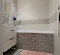 Fürdőszoba káddal, szürke csempékkel és rózsaszín szőnyeggel egy háromszobás lakásban.